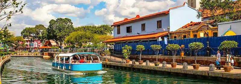 Ein blaues Boot fährt auf einem ruhigen Kanal in der Stadt Malakka, umgeben von Bäumen und charmanten Gebäuden.