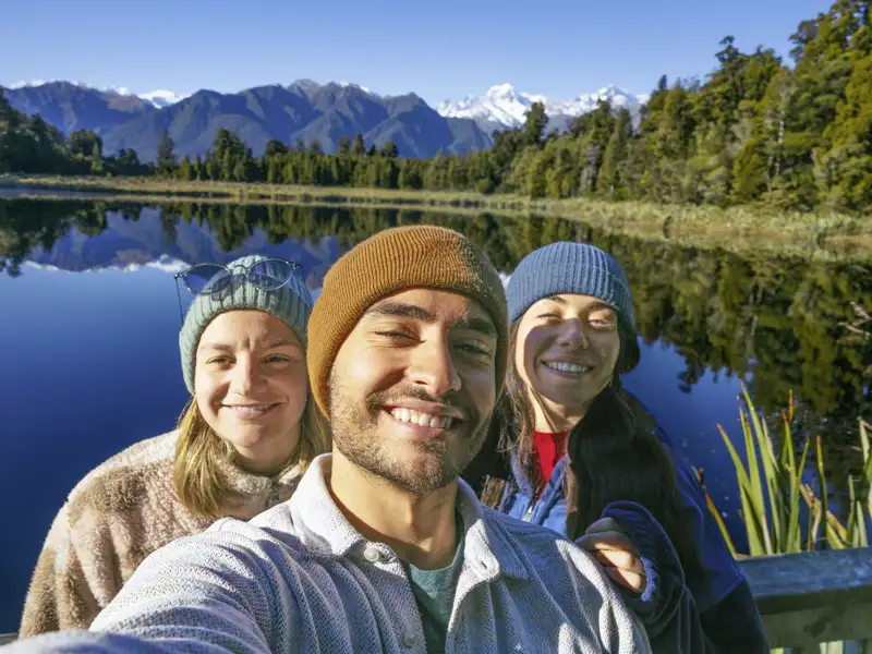 Drei junge Menschen machen ein Selfie vor einem See.
