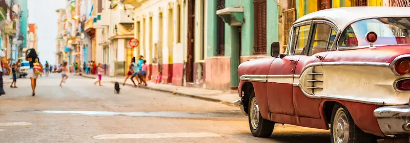 Klassischer Oldtimer steht am Straßenrand in Havanna vor bunten Häuserfassaden und spielenden Kindern.