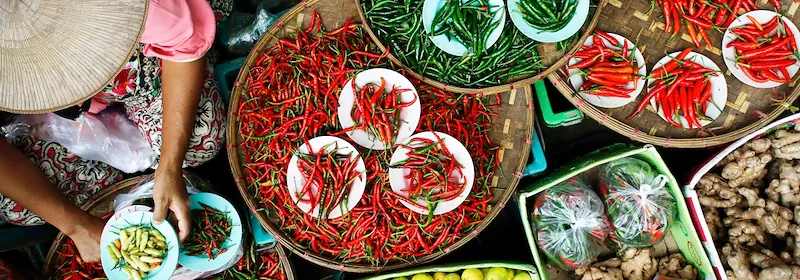 Marktszene in Laos von oben, bei der ein Händler roten und grünen Chilischoten, Gewürzen und Ingwer sortiert.