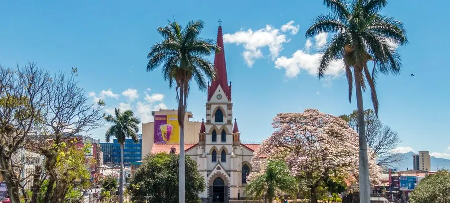 Frontalansicht der Kirche Iglesia de Nuestra Señora de la Merced im Stadtpark von San José, umgeben von Bäumen und Palmen bei sonnigem Wetter mit blauem Himmel.