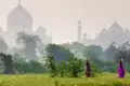 Zwei indische Personen in bunten Saris tragen Krüge auf dem Kopf durch ein grünes Feld, während sich das Taj Mahal im nebligen Hintergrund erhebt.