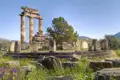 Blick auf die Überreste des Tholos-Tempels in Delphi vor wolkenfreiem Himmel. Rund um den Tempel wächst das Gras hoch und auch einige bunte Blumen sind zu sehen.