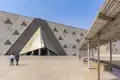 Besucher spazieren über den sonnigen Vorplatz auf den pyramidenförmigen Eingang des Grand Egyptian Museum in Gizeh zu.