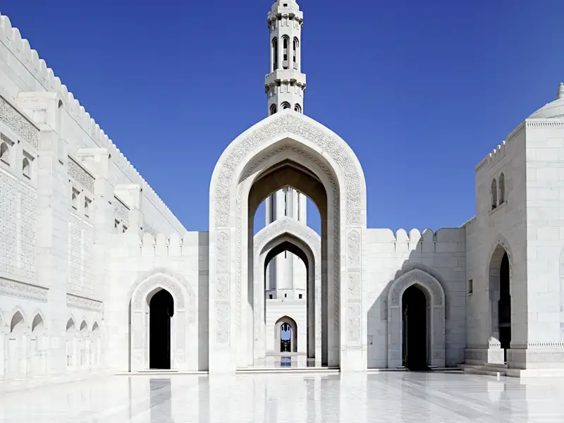 Blick auf einen der Innenhöfe der Sultan-Qaboos-Moschee in Maskat mit weiß glänzendem Marmorboden. Durch einen spitzen Torbogen erblickt man einen Teil des Minaretts.