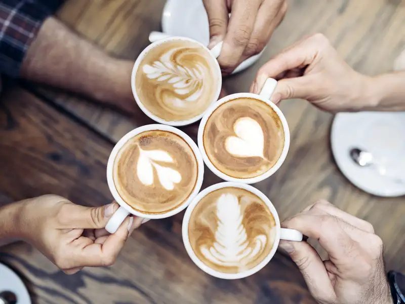 Hände einer jungen Reisegruppe halten vier Tassen Cappuccino mit kunstvoller Latte Art über einem Holztisch.