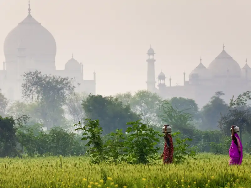 Zwei indische Personen in bunten Saris tragen Krüge auf dem Kopf durch ein grünes Feld, während sich das Taj Mahal im nebligen Hintergrund erhebt.