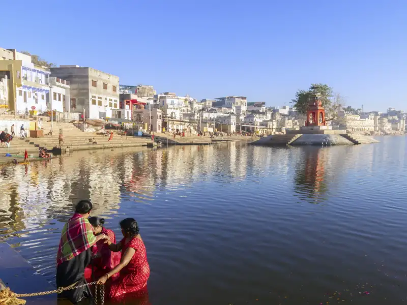 Drei Personen in farbenfrohen Saris bei einer rituellen Waschung im heiligen Pushkar-See, einem Highlight der Rajasthan-Reise, mit den Ghats und der Stadt Pushkar im Hintergrund.