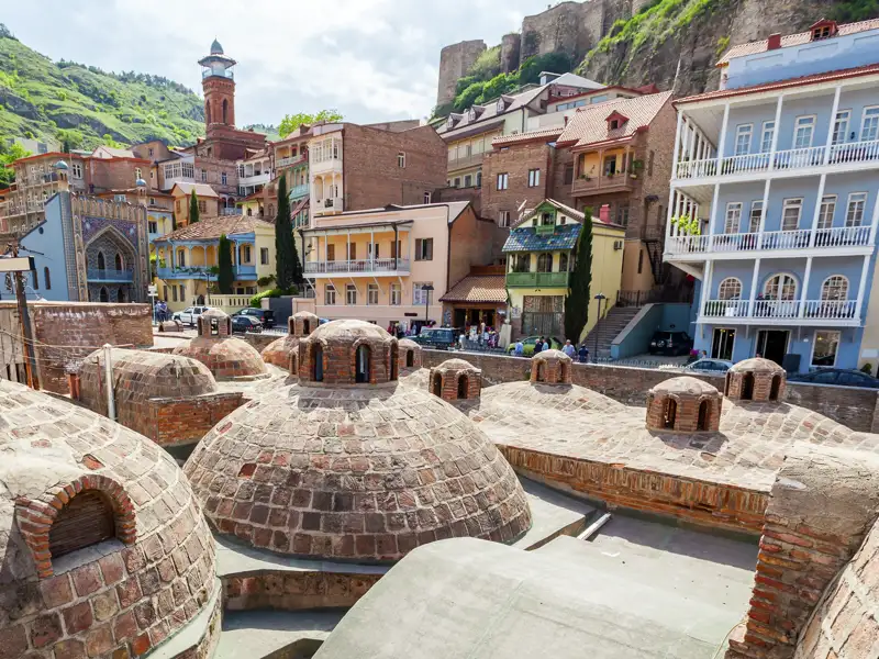 Blick über die historischen Kuppeldächer der Schwefelbäder auf die bunte Altstadt von Tiflis und die Nariqala-Festung.