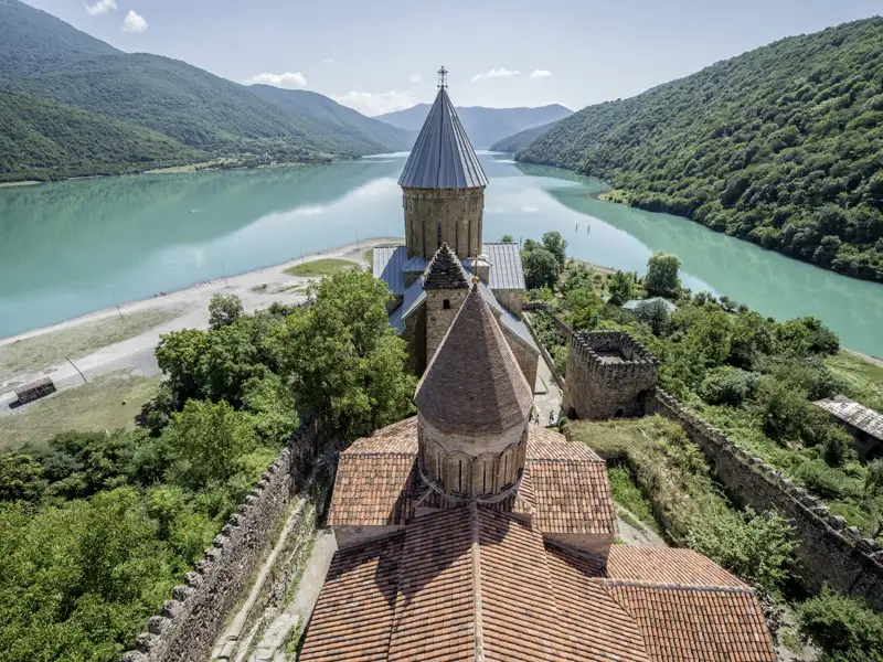 Die historische Wehrkirche Ananuri in Georgien am Ufer des türkisgrünen Schinwali-Stausees vor Kaukasus-Bergkulisse.