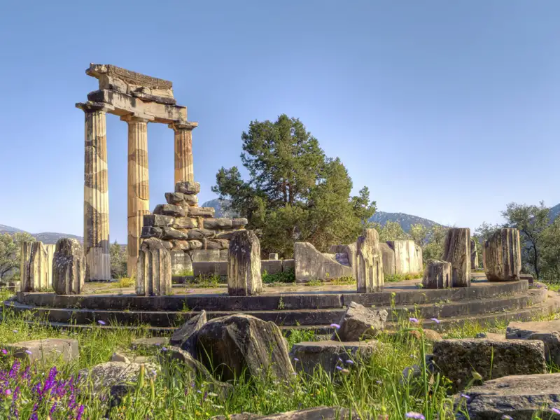 Blick auf die Überreste des Tholos-Tempels in Delphi vor wolkenfreiem Himmel. Rund um den Tempel wächst das Gras hoch und auch einige bunte Blumen sind zu sehen.