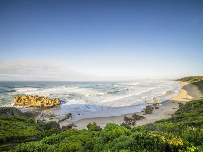 Weite Küstenlandschaft an Südafrikas Gartenroute mit Sandstrand, Wellen, Felsen und üppig grüner Vegetation.