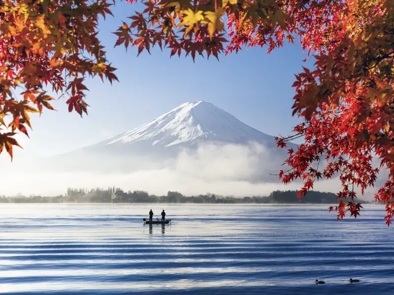 Zwei Personen in einem kleinen Boot auf einem See vor dem schneebedeckten Gipfel des Fuji-san, umrahmt von rotem Herbstlaub.