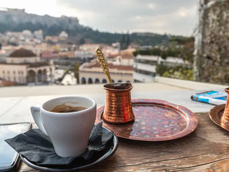 Eine Tasse Kaffee und ein traditionelles Briki-Kännchen auf einem Holztisch mit malerischem Blick auf die Akropolis in Athen.