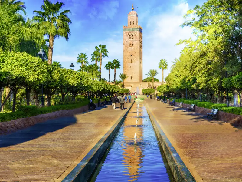 Langer Wasserkanal im Park vor der Koutoubia-Moschee in Marrakesch, gesäumt von Palmen, in dem sich das Minarett spiegelt.