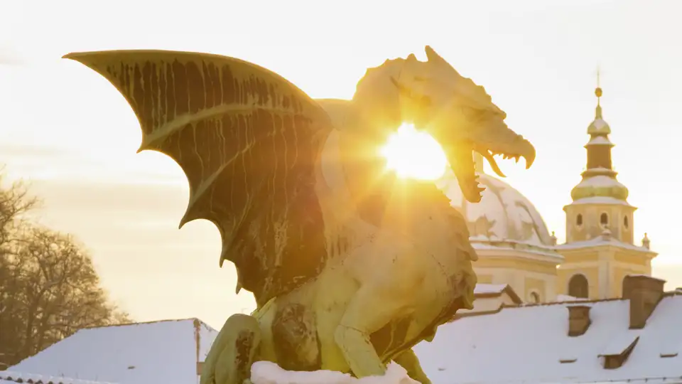 Die Drachenstatue auf der Drachenbrücke in Ljubljana im Gegenlicht der Wintersonne, im Hintergrund schneebedeckte Dächer.