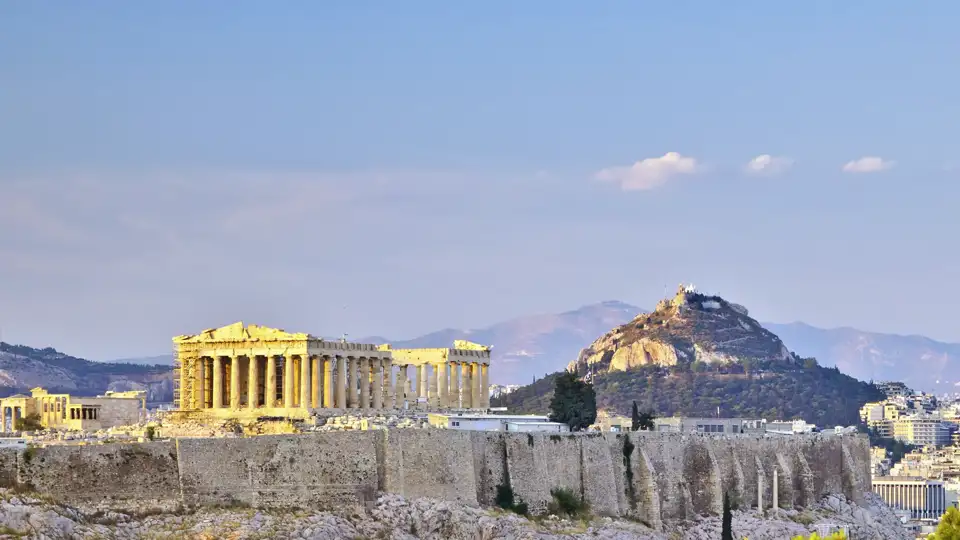 Blick auf den Parthenon-Tempel auf dem Felsen der Akropolis in Athen mit dem markanten Lykabettus-Hügel im Hintergrund.